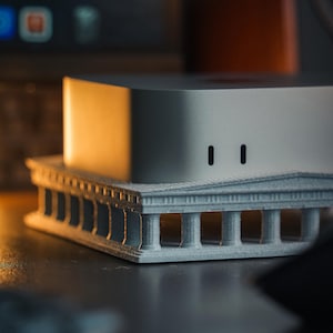 Puede incluir: Un dispositivo electrónico de forma cuadrada, color plateado, con esquinas redondeadas y dos ranuras verticales negras en la parte frontal, apoyado sobre una base arquitectónica gris claro impresa en 3D con columnas y un frontón.
