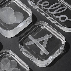 Puede incluir: Una colección de bloques de acrílico transparente con diseños grabados. Un bloque rectangular muestra la palabra hello en letra cursiva. Otros bloques cuadrados presentan gráficos grabados que incluyen una letra A mayúscula, un patrón floral y formas geométricas.