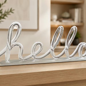 May include: A clear glass 'hello' sign in a cursive font, mounted on a silver base. The sign is displayed on a light brown wooden surface. A vase with greenery is in the background. The sign is a decorative item.