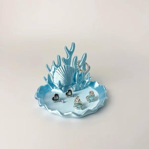 May include: A light blue jewellery dish featuring a sculpted coral and seashell design. The base is a scalloped tray holding four small rings with heart-shaped and round stones, plus one beaded ring resting on the upright coral structure.