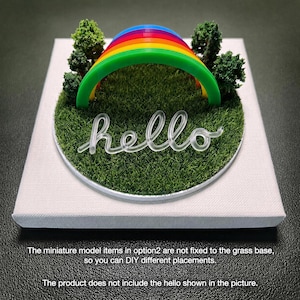 Op de afbeelding: Een miniatuur diorama met een veelkleurige regenboogboog en kleine groene bomen op een cirkelvormig stuk kunstgras. Het geheel is gemonteerd op een vierkante witte canvas basis. De tekst hello is op het gras geplaatst.