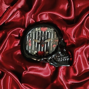 May include: A black skull-shaped decorative object with an oval-shaped interior featuring a black and white striped background, flowers, and a bee. The object is set against a backdrop of red satin fabric.