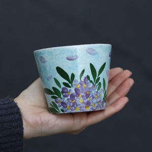 May include: A small, hand-painted ceramic pot with a light blue background and a floral design. The pot features purple flowers, green leaves, and small purple cloud-like accents. The pot is held in a person's hand against a dark background.