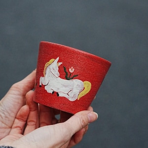 May include: A red, cylindrical planter with a white horse illustration and a yellow mane and tail. The planter also features a small red flower. The planter is held in two hands against a gray background.