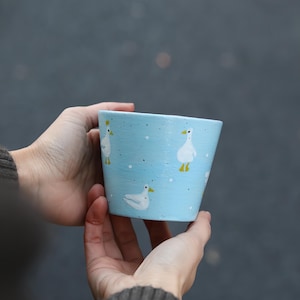 May include: A light blue tapered plant pot featuring a hand-painted pattern of white geese with yellow beaks and feet. The surface of the pot is decorated with small white dots and black speckles, creating a simple, repeating motif across the exterior.