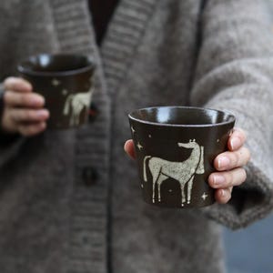 May include: Two dark brown ceramic mugs with a white horse design and star accents are held by a person wearing a gray cardigan. The mugs have a glossy finish and a simple, rustic aesthetic, perfect for coffee or tea.