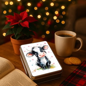 May include: A metal tin featuring a watercolour illustration of a black and white cow with pink hearts. A cup of coffee, biscuits, and a poinsettia plant are also visible on a wooden table.