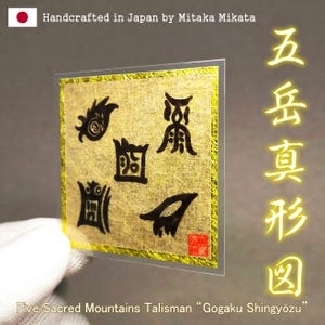 Puede incluir: Un talismán dorado con símbolos y texto en negro, enmarcado en plástico transparente. El texto dice "Five Sacred Mountains Talisman Gogaku Shingyōzu" y "Handcrafted in Japan by Mitaka Mikata."