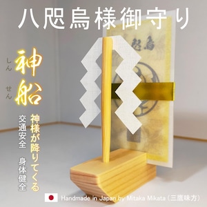 May include: A wooden Japanese Shinto shrine ornament. The ornament features a light-colored wooden base and a vertical yellow pole with a white zigzag paper design. The background includes a wrapped paper item with Japanese text.