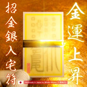 Puede incluir: Un amuleto japonés rectangular, amarillo y blanco con detalles dorados y caracteres japoneses. El amuleto está hecho a mano en Japón. El texto incluye "招金銀人宅待" y "金運上昇".