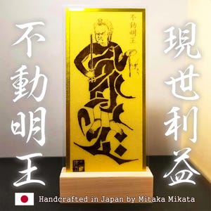 Puede incluir: Una obra de arte dorada con una ilustración negra de una figura sosteniendo una espada y una cuerda. Hay caracteres japoneses a la izquierda y a la derecha. La obra está montada sobre una base de madera. El texto en la parte inferior dice "Handcrafted in Japan by Mitaka Mikata."