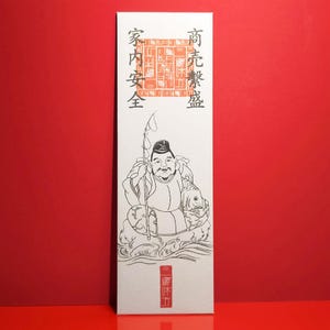 May include: A rectangular white paper with a Japanese illustration of a seated figure holding a staff and a fish. The illustration is in black ink with red accents. Japanese text is printed at the top and bottom of the paper.