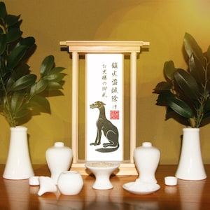 May include: A Japanese Shinto altar featuring a wooden frame and a scroll with a black dog illustration and Japanese text. White ceramic vases and bowls are arranged around the altar, with green foliage and a wooden table.