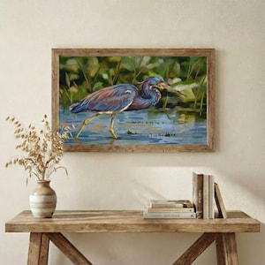 May include: A framed painting depicts a heron wading in water. The bird has blue, brown, and white feathers, with a long yellow beak and legs. The background features green foliage and water, with a wooden frame.