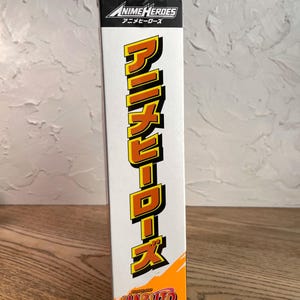 May include: A white box featuring the text "ANIME HEROES" in a black and white logo. Below, the Japanese characters for "Anime Heroes" are printed. The box also displays the Japanese text "アニメヒーローズ" in a vertical, yellow font, and the "NARUTO Shippuden" logo.