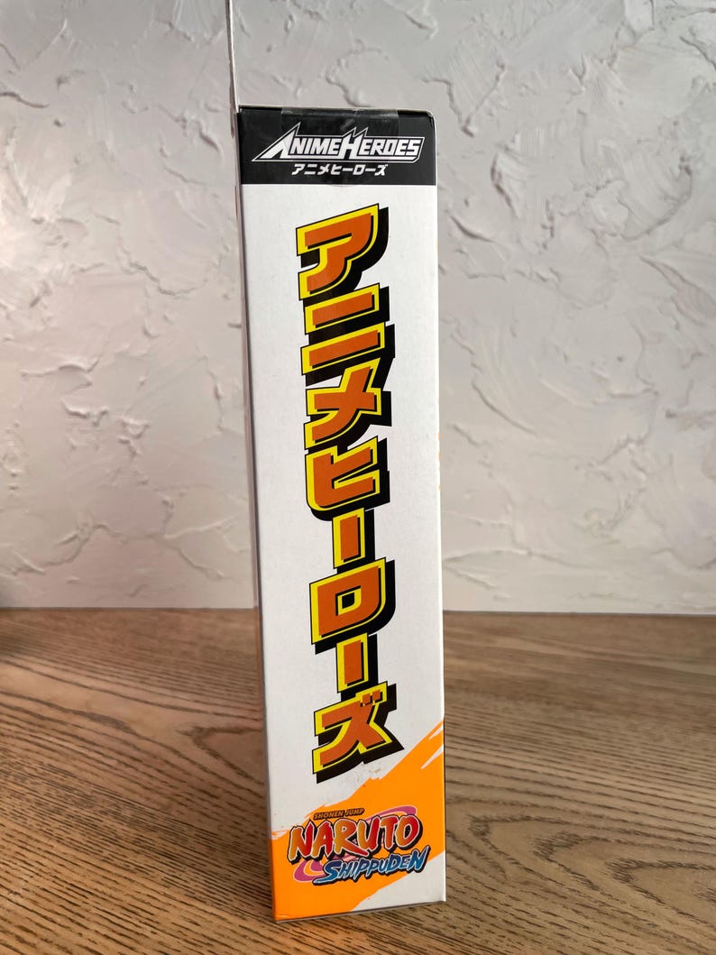 May include: A white box featuring the text "ANIME HEROES" in a black and white logo. Below, the Japanese characters for "Anime Heroes" are printed. The box also displays the Japanese text "アニメヒーローズ" in a vertical, yellow font, and the "NARUTO Shippuden" logo.