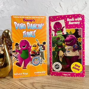 May include: Two vintage Barney VHS tapes. One tape is orange with the title "Dino Dancin' Tunes" and features Barney and a band. The other tape is pink and says "Rock with Barney". Both tapes are sealed.