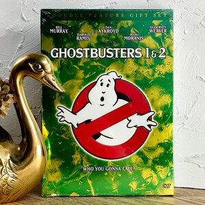 以下が含まれることがあります： 映画「ゴーストバスターズ1&2」のDVDギフトセット。表紙は緑色で、ゴーストバスターズのロゴと「Who You Gonna Call?」の文字が入っています。出演者はビル・マーレイ、ダン・エイクロイド、シガニー・ウィーバーなど。金色の白鳥の置物が前景にあります。