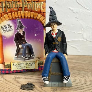 May include: A Harry Potter collectible figurine of Harry Potter wearing a black wizard hat and glasses, seated on a stone-like base. He wears a black jacket, red and white striped shirt, and blue jeans. The figurine is next to a Harry Potter box.