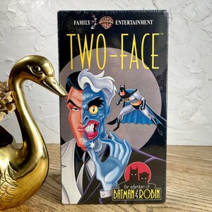 May include: A vintage VHS tape for "Two-Face" from "The Adventures of Batman & Robin." The cover features a split image of Two-Face and Batman in action, with the Warner Bros. Family Entertainment logo at the top.