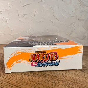 May include: A white box with the words "Naruto Shippuden" in orange, red, and blue. The box has a black and orange design on the top and the words "Shonen Jump" at the top.
