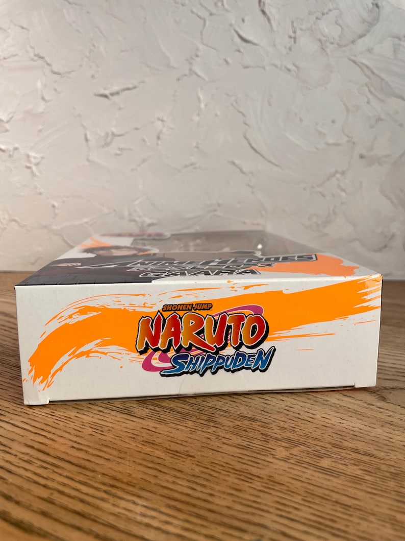 May include: A white box with the words "Naruto Shippuden" in orange, red, and blue. The box has a black and orange design on the top and the words "Shonen Jump" at the top.