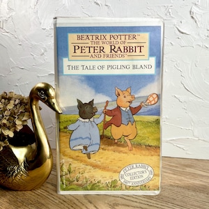 May include: A vintage VHS tape of "The Tale of Pigling Bland" from the Peter Rabbit and Friends series. The cover features illustrations of two pigs in a pastoral setting. The tape is a collector's edition celebrating the 100th anniversary.