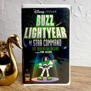 May include: A VHS tape of Disney Pixar's "Buzz Lightyear of Star Command: The Adventure Begins" featuring Tim Allen. The cover has a green and white title with a space-themed background and a cartoon image of Buzz Lightyear.