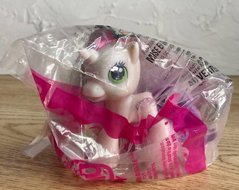 May include: A light pink plastic unicorn toy figure with a purple mane and tail, featuring large green eyes and a small horn on its forehead. The toy is sealed inside a clear plastic bag with pink accents and printed safety warning text.