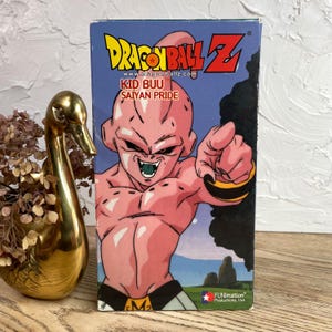 May include: A Dragon Ball Z VHS tape featuring Kid Buu. The box has a blue background with the Dragon Ball Z logo and the text "Kid Buu Saiyan Pride." The character Kid Buu is pink and pointing. The box is next to a gold swan figurine.