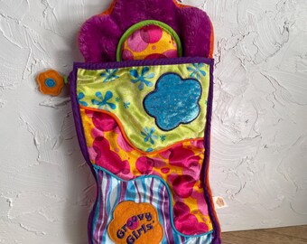 May include: A plush sleeping bag for dolls featuring a purple flower-shaped top, a lime green section with blue flower accents, and a lower portion with pink, orange, and blue striped patterns. An orange patch with the text Groovy Girls is attached.