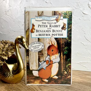 May include: A vintage book titled "The Tale of Peter Rabbit and Benjamin Bunny" by Beatrix Potter. The cover features an illustration of Peter Rabbit. The book is a collector's edition celebrating its 100th anniversary.