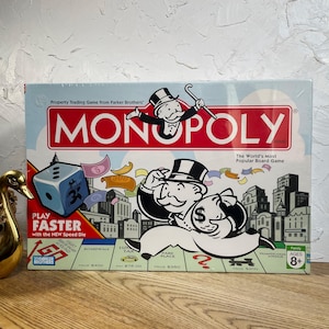 May include: A Monopoly board game box featuring the iconic character Rich Uncle Pennybags in a top hat, a large blue die, and colorful play money. The box displays the text Monopoly, Play Faster with the New Speed Die, and The World's Most Popular Board Game.