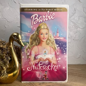 May include: A VHS tape for "Barbie in the Nutcracker" movie. The cover features Barbie with blonde hair and a tiara, wearing a pink dress. The title is in a cursive font, with a pink and purple background.