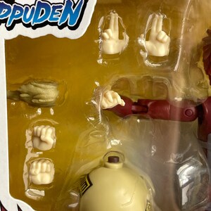 May include: A packaged action figure from the Naruto Shippuden series. The figure is red and tan with multiple interchangeable hand gestures and a yellow spherical accessory. The packaging includes the series logo in red, white, and blue.