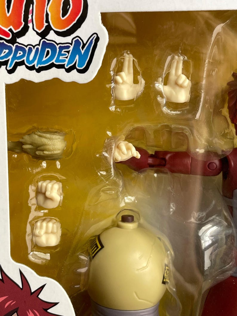 May include: A packaged action figure from the Naruto Shippuden series. The figure is red and tan with multiple interchangeable hand gestures and a yellow spherical accessory. The packaging includes the series logo in red, white, and blue.