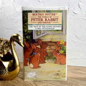 May include: A vintage VHS tape of "The World of Peter Rabbit and Friends" by Beatrix Potter. The cover features illustrations of brown bunnies and the text "The Tale of the Flopsy Bunnies & Mrs. Tittlemouse". A gold swan figurine is in the foreground.