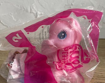 May include: A pink My Little Pony toy figure with a long mane and tail, enclosed in a clear plastic bag with pink accents. The packaging features the text My Little Pony and the number 2 printed in white on a pink background, visible through the plastic.