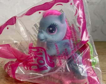 May include: A light blue My Little Pony toy figure with bright pink hair, large purple eyes, and a small heart detail, sealed inside a clear plastic bag with pink trim and printed text including the name Rainbow Dash and safety warnings.