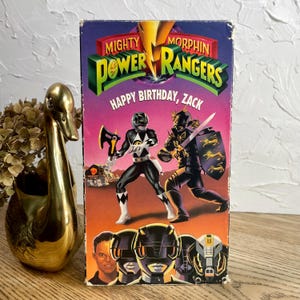 May include: A vintage VHS tape for "Mighty Morphin Power Rangers" with the text "Happy Birthday, Zack." The cover features illustrations of Power Rangers in action poses, with a gold and purple color scheme.