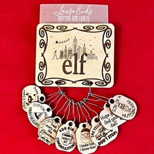 May include: A collection of wooden knitting stitch markers and a wooden sign. The sign features the word "elf" with a skyline design. The stitch markers have various engraved phrases and images, including "Cotton-Head" and "Son of a Nutcracker!"