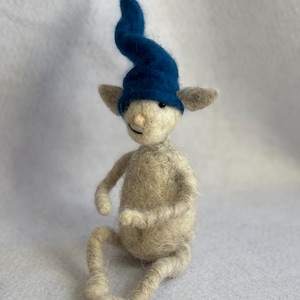 May include: A handmade felted gnome figurine. The gnome has a light beige body, grey ears, and a long, curved blue hat. It is sitting with its legs extended and arms bent. A whimsical design.