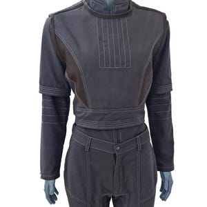 Op de afbeelding: Een donkergrijze tweedelige outfit met een hoge kraag en lange mouwen. De top heeft verticale stiksels en een riem. De broek heeft knielappen en een knoopsluiting. De outfit wordt op een mannequin getoond.