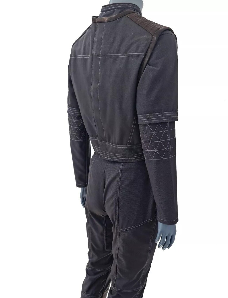 Op de afbeelding: Een donkergrijze jumpsuit met witte stikseldetails. De jumpsuit heeft een hoge kraag, schouderstukken en geometrische patronen op de mouwen. Het kledingstuk wordt op een mannequin getoond.