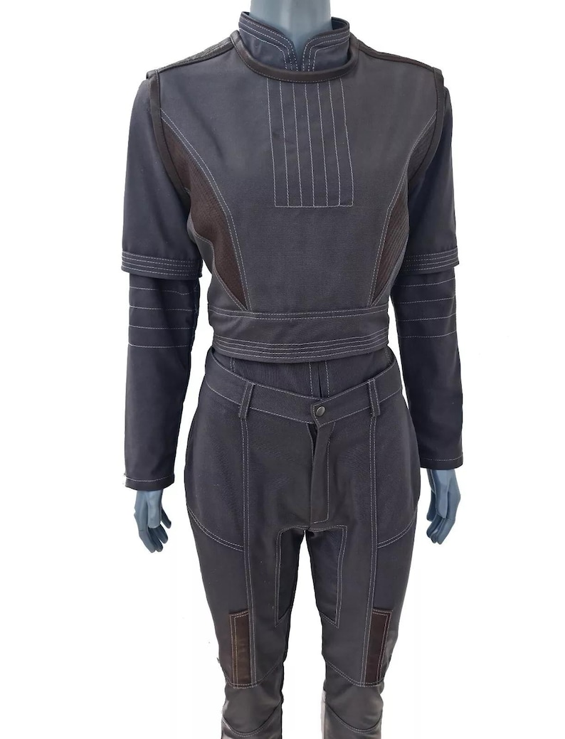 Op de afbeelding: Een donkergrijze tweedelige outfit met een hoge kraag en lange mouwen. De top heeft verticale stiksels en een riem. De broek heeft knielappen en een knoopsluiting. De outfit wordt op een mannequin getoond.