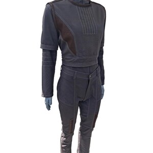 Op de afbeelding: Een donkergrijze jumpsuit met bruine accenten en witte stiksels. De top heeft een hoge kraag en korte mouwen met een wikkelriem. De broek is voorzien van knielappen. De outfit wordt getoond op een mannequin.