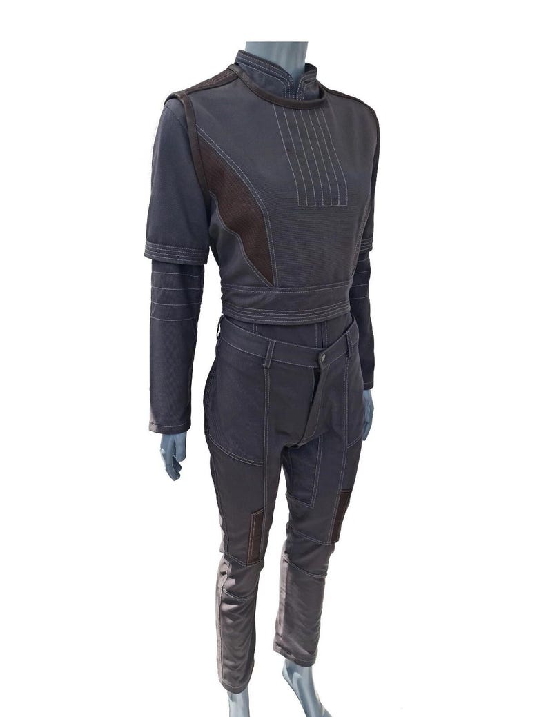 Op de afbeelding: Een donkergrijze jumpsuit met bruine accenten en witte stiksels. De top heeft een hoge kraag en korte mouwen met een wikkelriem. De broek is voorzien van knielappen. De outfit wordt getoond op een mannequin.