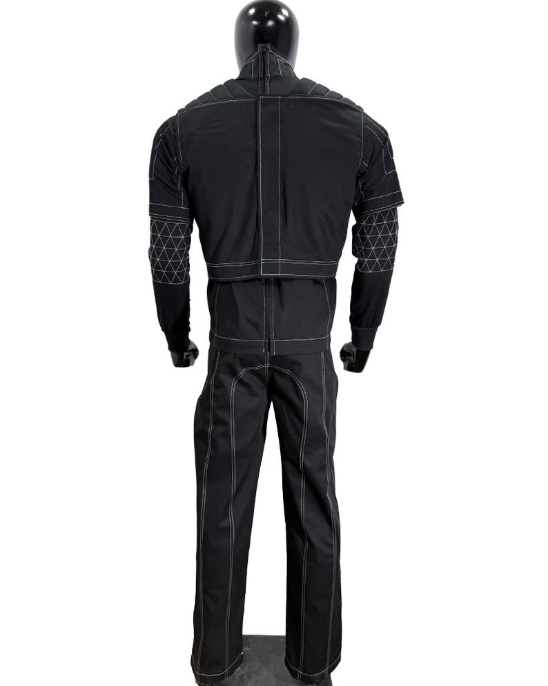 May include: Black jumpsuit with white stitching and geometric patterned accents on the sleeves. The outfit is displayed on a mannequin. The jumpsuit has a high collar and long sleeves. The pants are straight-legged.