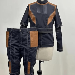 Puede incluir: Un conjunto de dos piezas azul marino, compuesto por una chaqueta y un pantalón. La chaqueta tiene detalles marrones en los hombros y el pecho, con costuras blancas. El pantalón tiene bolsillos marrones y costuras blancas. El conjunto se exhibe en un maniquí.