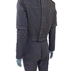 Op de afbeelding: Een donkergrijze jumpsuit met bruine schouderaccenten en witte stiksels. De mouwen hebben geometrische patronen bij de manchetten. De achterkant heeft verticale en horizontale stikseldetails.